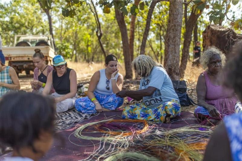A lady weaves under the shade of a tree with others around watching