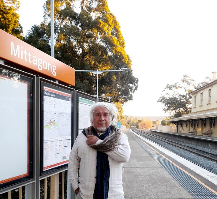 The artist Djon Mundine stands in front of a train station sign saying 'Mittagong', with the sun setting in the background.